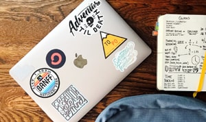 What Is a Sticker? The Difference Between Stickers article preview with MacBook with stickers and notebook.